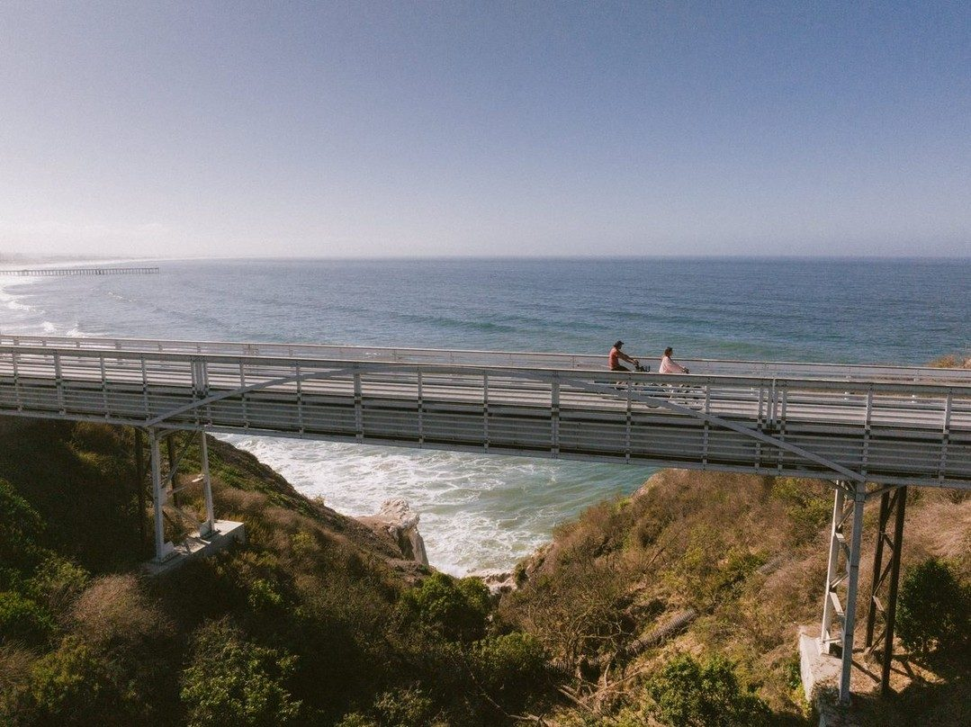 Two people ride bikes across a bridge overlooking the ocean, with waves crashing on rocks below and greenery on the cliffs. The sky is clear and blue, and the horizon stretches into the distance.