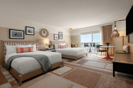 A modern hotel room with two queen beds, wall art, a round table with chairs, a bench, and a large window with curtains leading to a balcony overlooking the ocean.