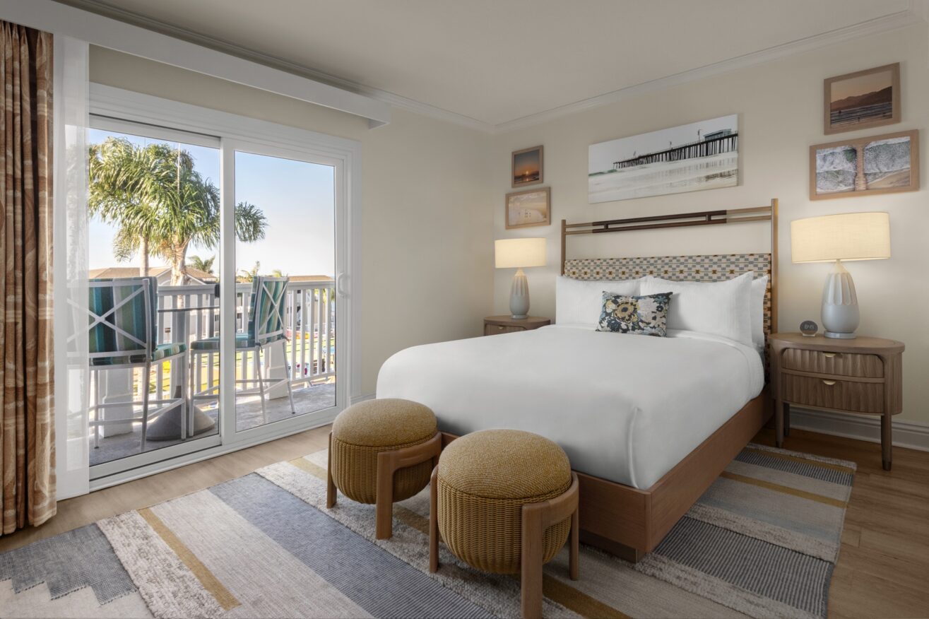 A bright hotel room with a neatly made bed, two round ottomans at the foot, nightstands with lamps, coastal artwork on the walls, and a sliding glass door leading to a balcony with palm trees outside.