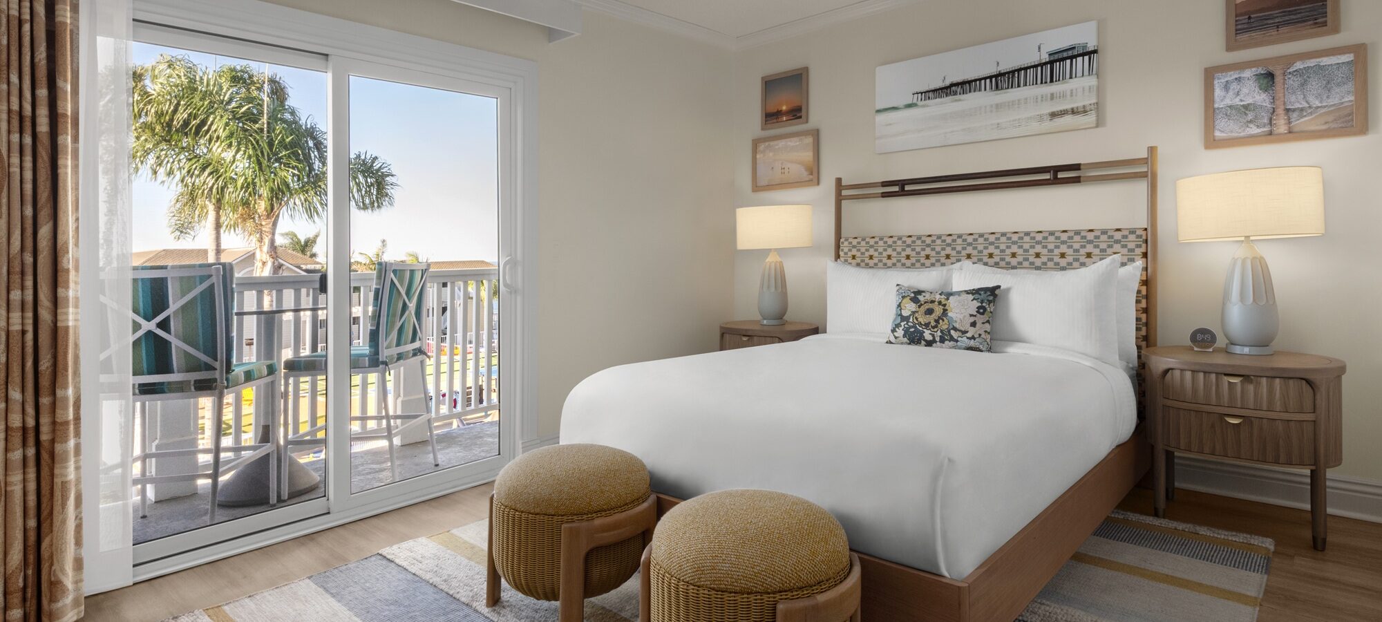 A bright hotel room with a neatly made bed, two round ottomans at the foot, nightstands with lamps, coastal artwork on the walls, and a sliding glass door leading to a balcony with palm trees outside.