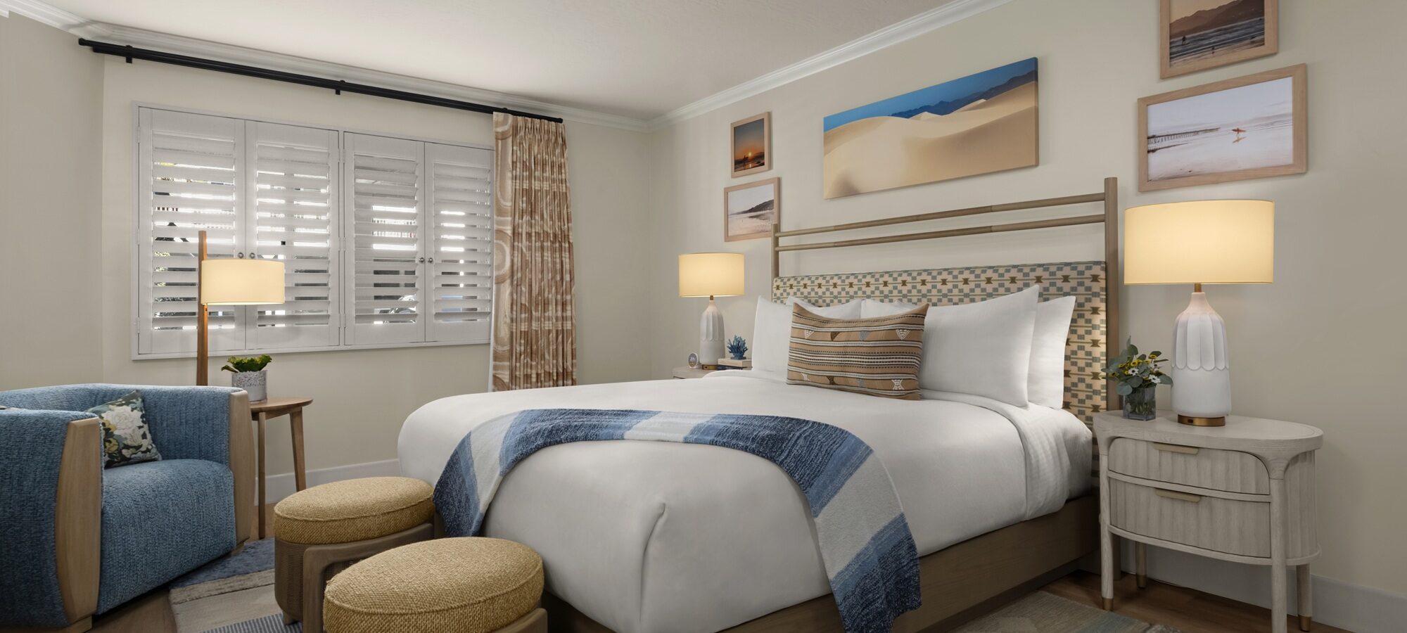 A cozy, modern bedroom with a large bed, blue and beige accents, two round ottomans, a blue armchair, two nightstands with lamps, wall art above the bed, and sunlight streaming through white shutters.
