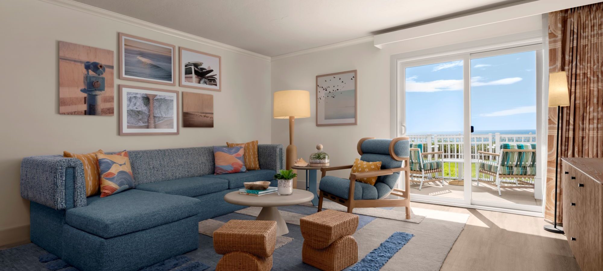 A bright living room with a blue sectional sofa, artwork on white walls, a round coffee table, armchair, woven stools, and a rug; sliding glass doors lead to a balcony with ocean views.