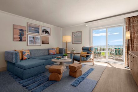 A bright living room with a blue sectional sofa, artwork on white walls, a round coffee table, armchair, woven stools, and a rug; sliding glass doors lead to a balcony with ocean views.