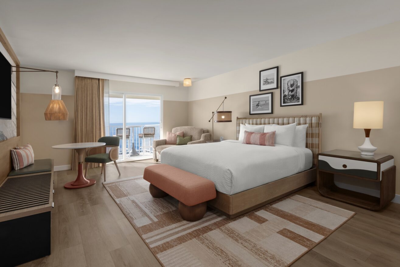A modern hotel room with a large bed, a nightstand, a lamp, a small dining table with two chairs, an armchair, and a balcony overlooking the ocean. The decor features warm, neutral tones and framed artwork.