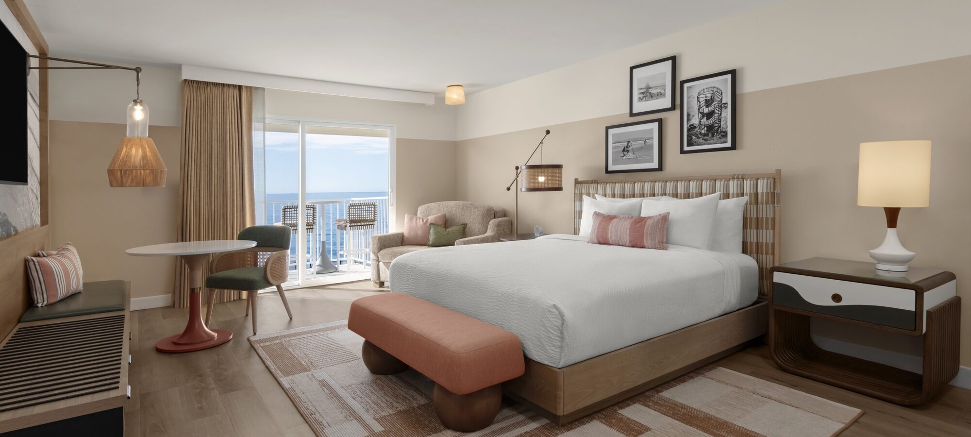 A modern hotel room with a large bed, a nightstand, a lamp, a small dining table with two chairs, an armchair, and a balcony overlooking the ocean. The decor features warm, neutral tones and framed artwork.