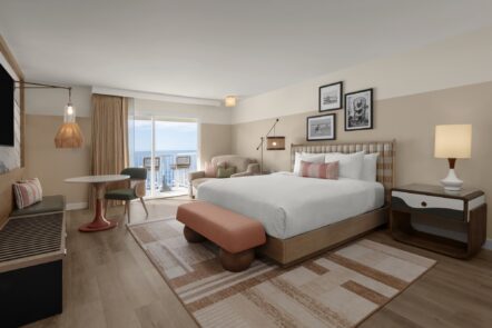 A modern hotel room with a large bed, a nightstand, a lamp, a small dining table with two chairs, an armchair, and a balcony overlooking the ocean. The decor features warm, neutral tones and framed artwork.