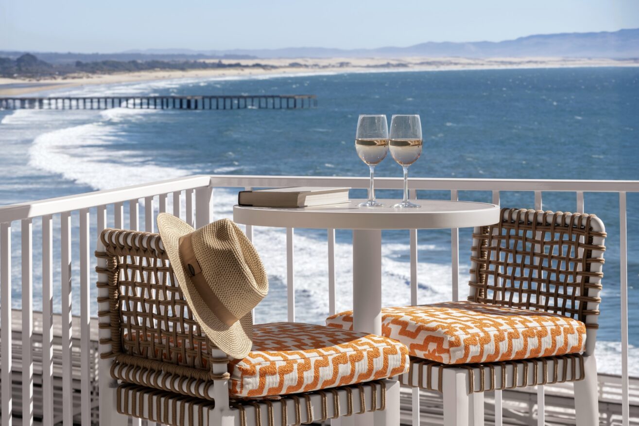 Two chairs with orange patterned cushions and a table with two glasses of white wine, books, and a straw hat overlook an ocean view with waves, a sandy beach, and a distant pier.