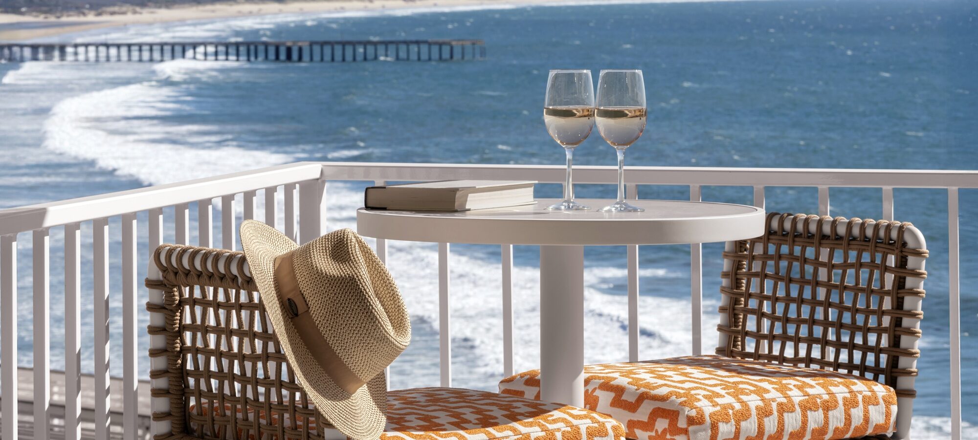 Two chairs with orange patterned cushions and a table with two glasses of white wine, books, and a straw hat overlook an ocean view with waves, a sandy beach, and a distant pier.