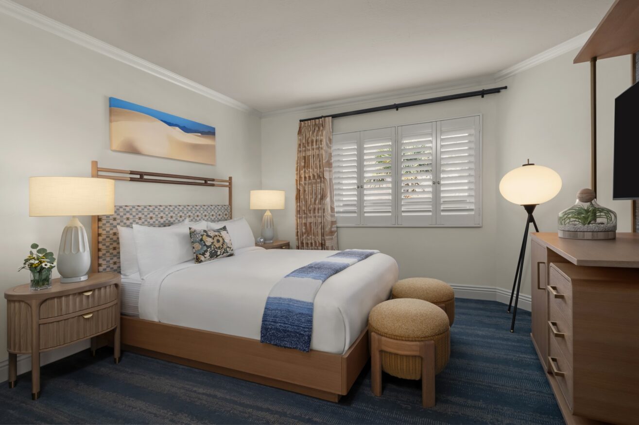 A cozy hotel room with a neatly made queen bed, wooden furniture, two round ottomans at the foot of the bed, blue accents, two bedside lamps, a globe floor lamp, a TV, and a window with white shutters.