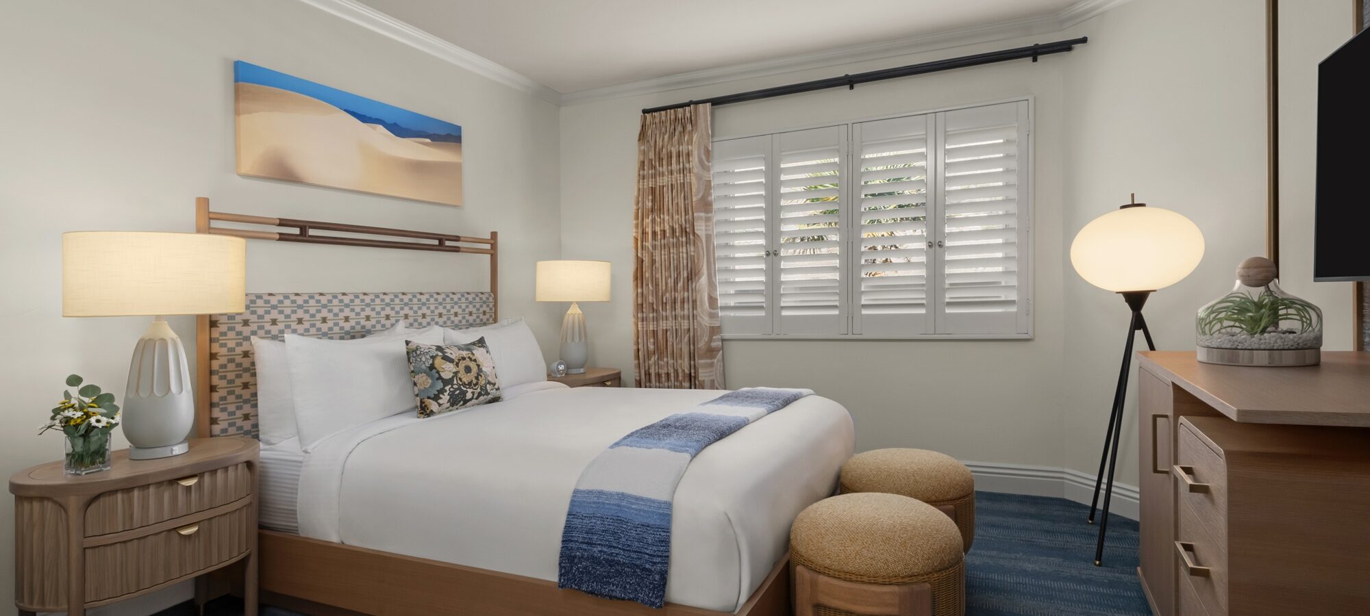 A cozy hotel room with a neatly made queen bed, wooden furniture, two round ottomans at the foot of the bed, blue accents, two bedside lamps, a globe floor lamp, a TV, and a window with white shutters.