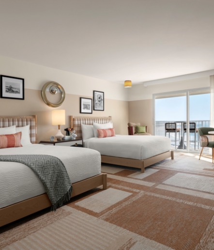 Modern hotel room with two double beds, patterned headboards, decorative pillows, and a blanket. There’s wall art, a lamp on a nightstand, and a balcony with chairs overlooking the ocean in the background.