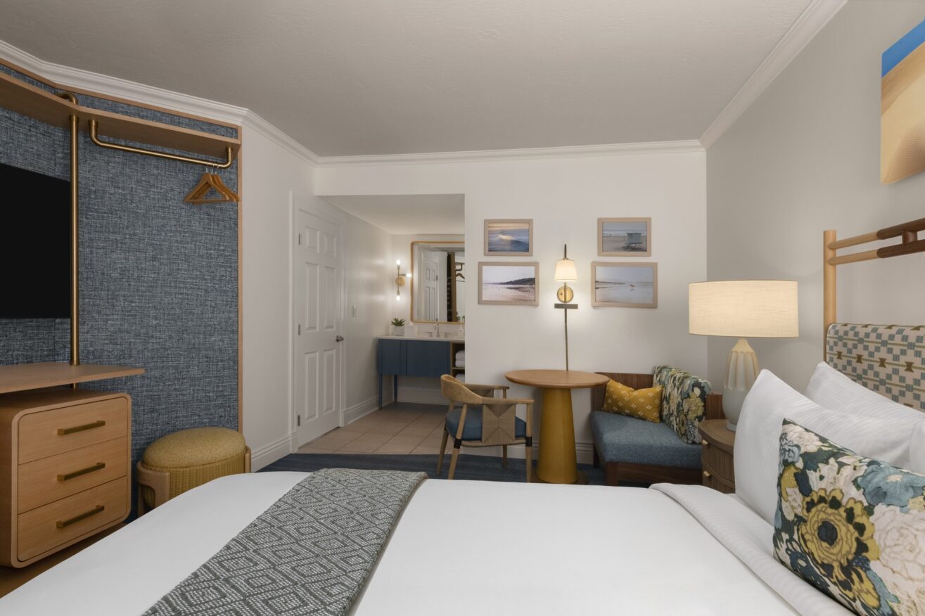 A neatly arranged hotel room with a bed in the foreground, a small round table with two chairs, a dresser, a TV, and an open closet. The bathroom is visible in the background, and there is wall art above the seating area.