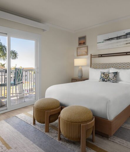 A cozy hotel room with a neatly made bed, two round ottomans, a nightstand with a lamp, artwork on the wall, and a balcony overlooking palm trees and a sunny outdoor scene through sliding glass doors.
