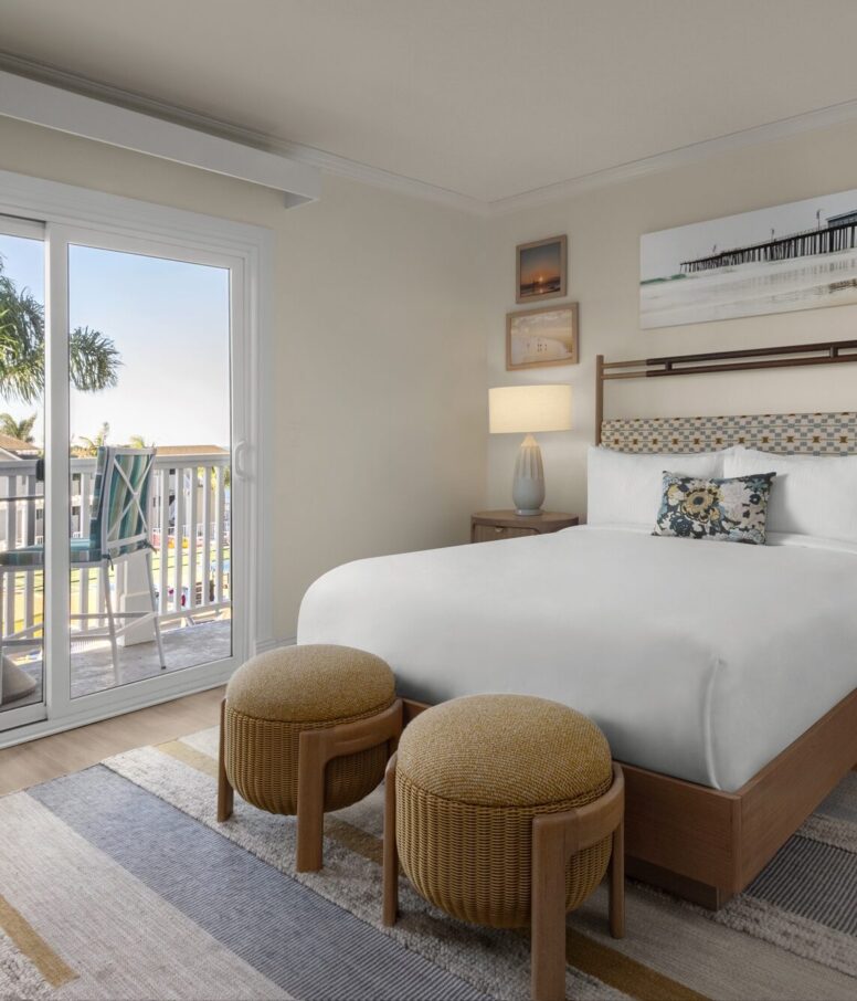 A cozy hotel room with a neatly made bed, two round ottomans, a nightstand with a lamp, artwork on the wall, and a balcony overlooking palm trees and a sunny outdoor scene through sliding glass doors.