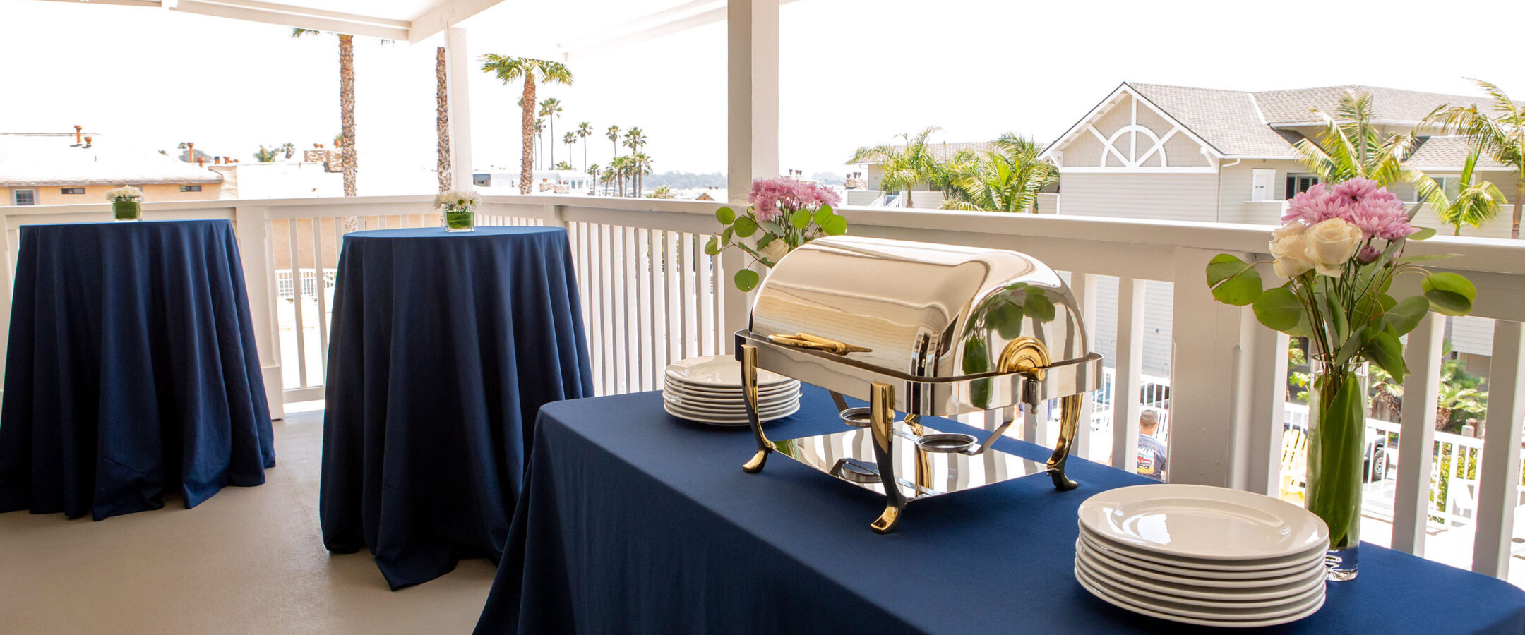 A covered outdoor patio setup with blue tablecloth-covered tables, a chafing dish, stacked white plates, and vases of flowers; palm trees and houses are visible in the background.