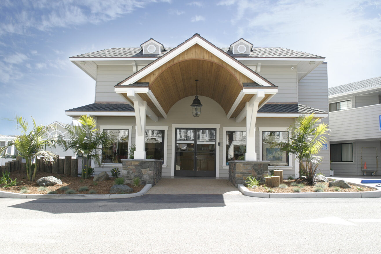 A modern two-story building with a large wooden entrance canopy, white siding, stone pillars, and landscaped plants in front, under a clear blue sky.