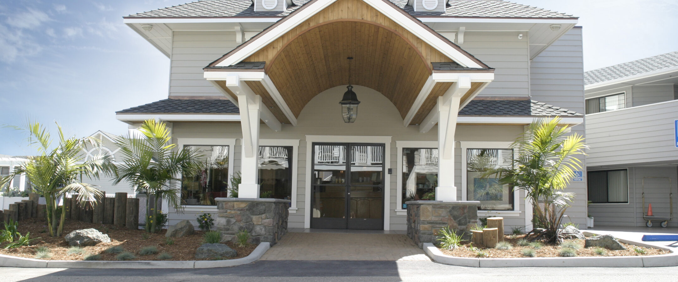 A modern two-story building with a large wooden entrance canopy, white siding, stone pillars, and landscaped plants in front, under a clear blue sky.