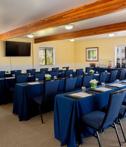 A conference room set up classroom-style with rows of tables covered in navy blue tablecloths, chairs, notepads, glasses, and small flower centerpieces. A TV screen and refreshment station are visible at the front.