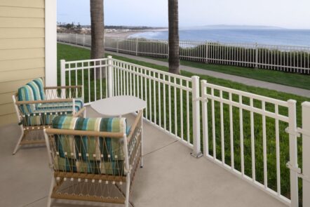 Two cushioned chairs and a small table sit on a patio with white fencing, overlooking a grassy area, palm trees, a coastal path, and the ocean in the background.
