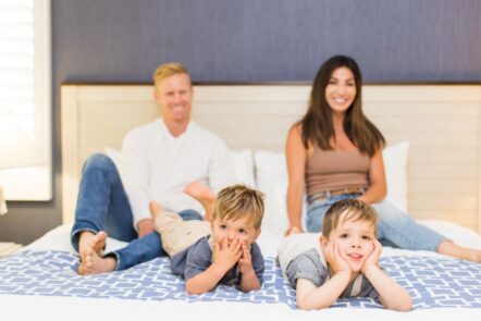 A smiling man and woman sit on a bed in the background, while two young boys lie at the foot of the bed in front, each resting their head on their hands and looking playful.