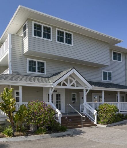 A modern, three-story building with light gray siding, white trim, and a covered porch with white railings. There are stairs leading to the entrance, and landscaped greenery surrounds the front.