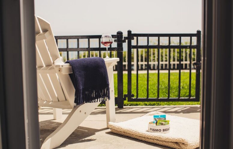 A white Adirondack chair with a navy throw blanket faces a black fence, overlooking a grassy area and ocean. A glass of red wine sits on the armrest; nearby, a basket labeled Pismo Beach holds snacks on a mat.