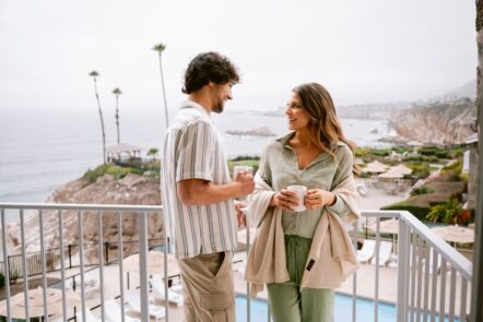 A man and woman stand on a balcony overlooking the ocean, both holding coffee mugs and smiling at each other. The background features palm trees, cliffs, and a cloudy sky.