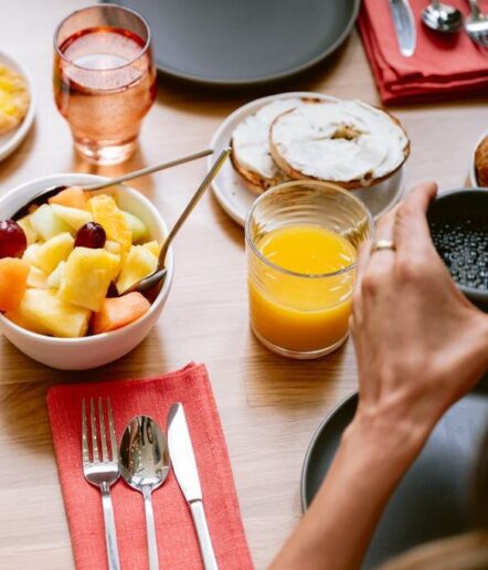A breakfast table with a bowl of mixed fruit, a glass of orange juice, a plate with a bagel and cream cheese, utensils on a red napkin, and a hand holding a cup of coffee.