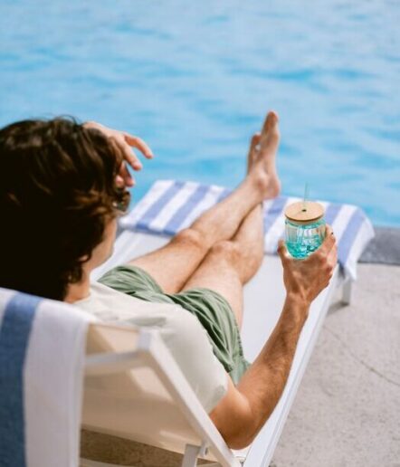 A person relaxing on a lounge chair by a pool, holding a blue drink with a straw and lid, with legs stretched out and a striped towel beneath them.
