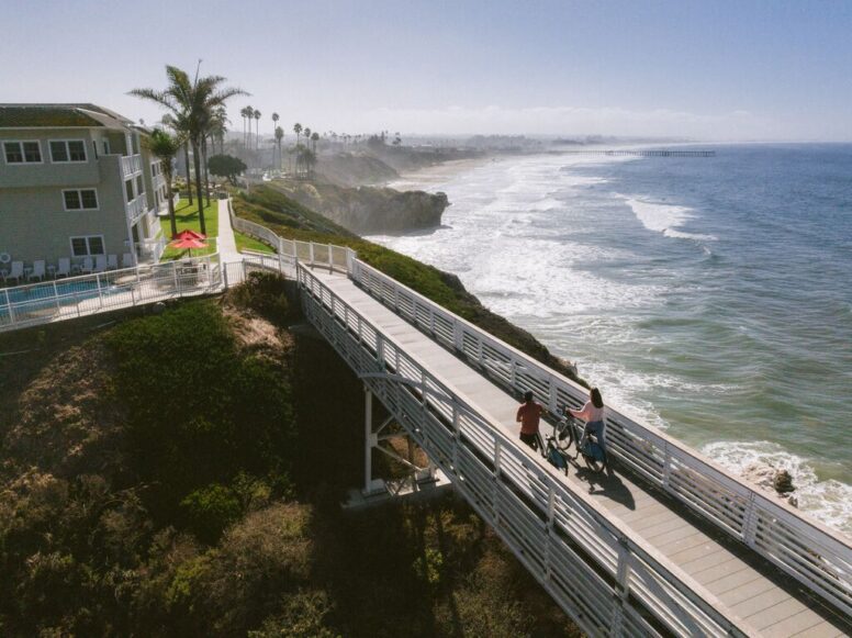 A man and a boy walk their bicycles along a coastal boardwalk overlooking the ocean, with waves crashing below and palm trees and houses on the cliffs in the background.