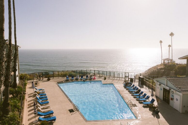 A rectangular outdoor swimming pool surrounded by blue lounge chairs overlooks the ocean at sunset, with a few palm trees, a small gazebo, and a clear sky in the background.