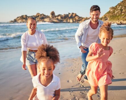 A smiling family of four, two adults and two young children, run and play together along a sunny beach with waves and rocky cliffs in the background. Everyone looks happy and carefree.