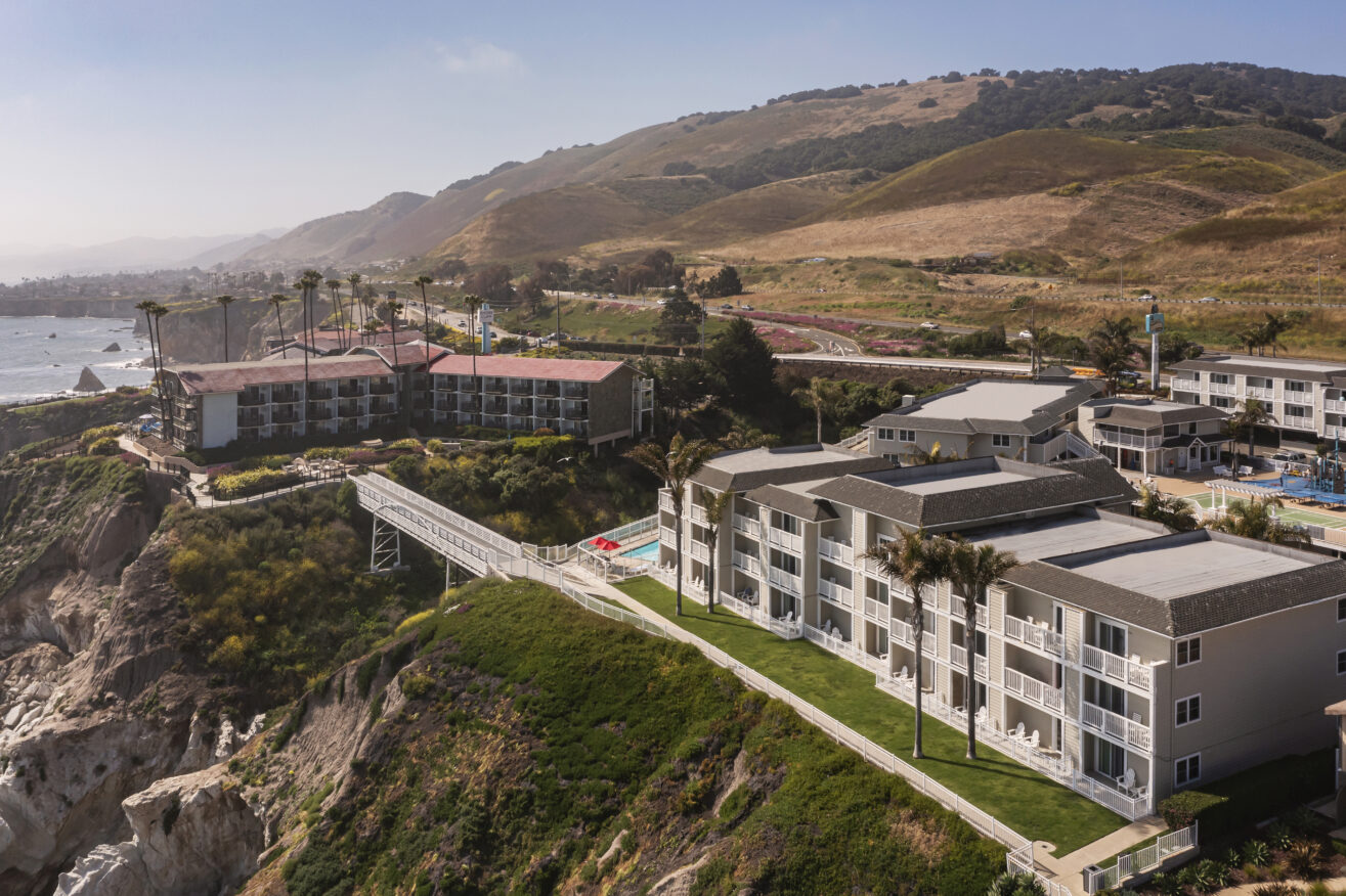 Aerial view of seaside resort buildings perched on a bluff overlooking the ocean, with hills and coastal landscape in the background. Palm trees and a walkway connect the buildings.