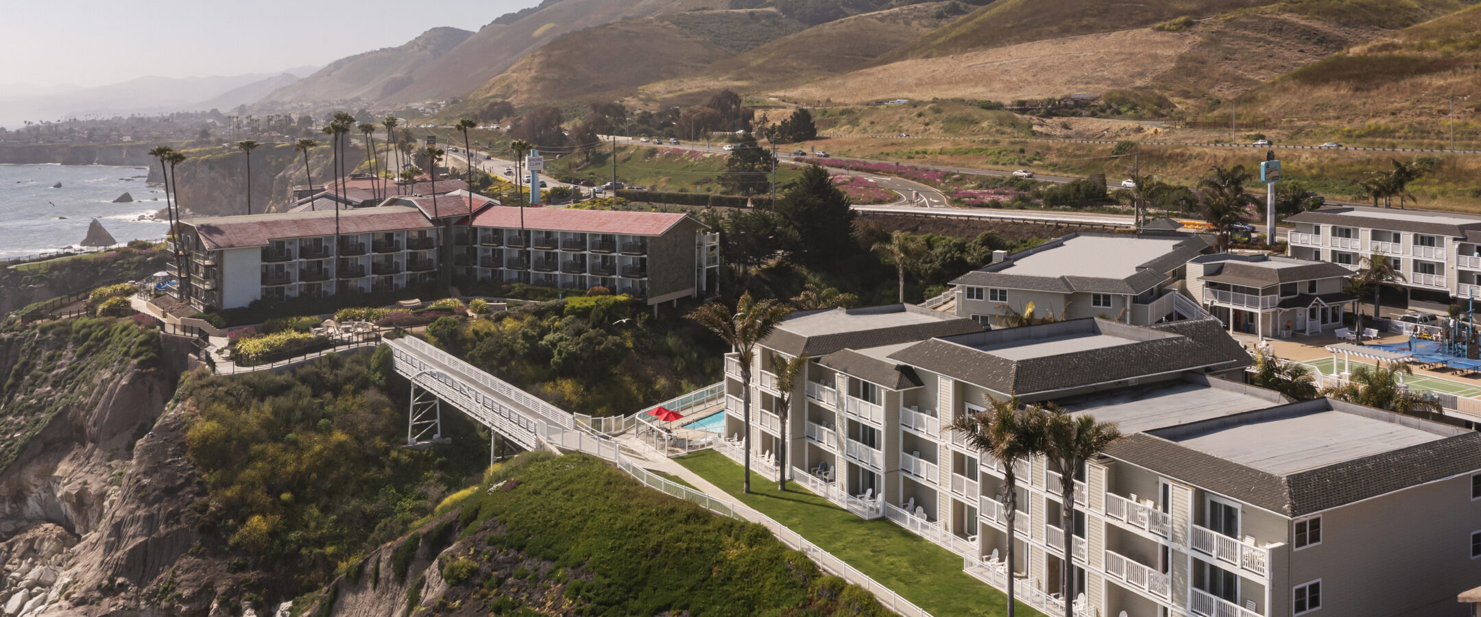 Aerial view of seaside resort buildings perched on a bluff overlooking the ocean, with hills and coastal landscape in the background. Palm trees and a walkway connect the buildings.