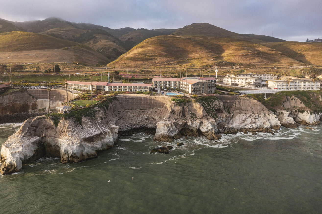 A coastal hotel complex sits atop rugged cliffs overlooking the ocean, with waves crashing below and rolling green hills in the background under a cloudy sky.