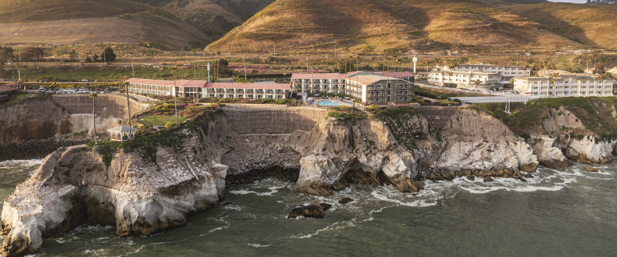 A coastal hotel complex sits atop rugged cliffs overlooking the ocean, with waves crashing below and rolling green hills in the background under a cloudy sky.