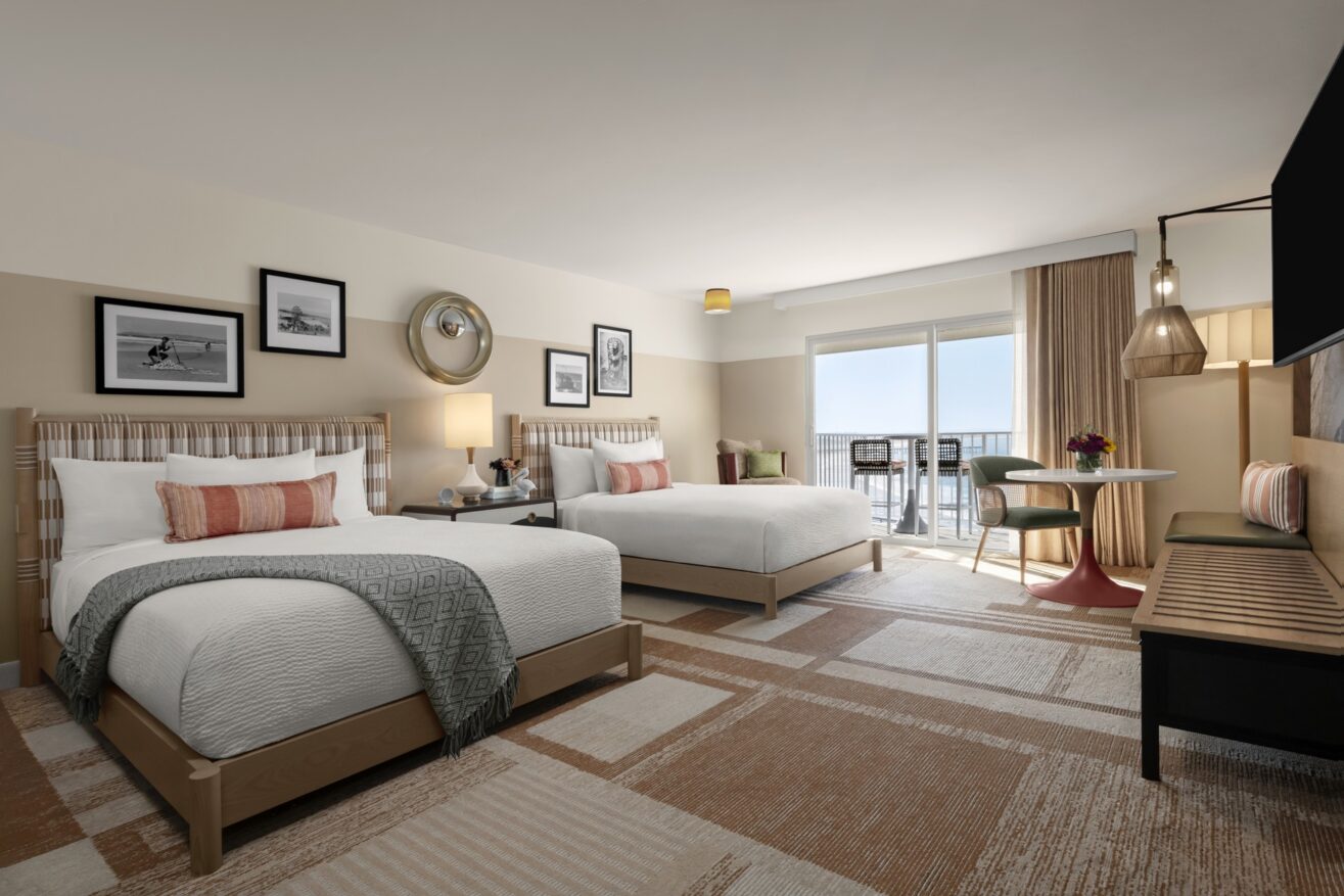 Modern hotel room with two double beds, decorative pillows, framed artwork on the walls, a round table with two chairs, a bench, and a large window with curtains leading to a balcony with an ocean view.