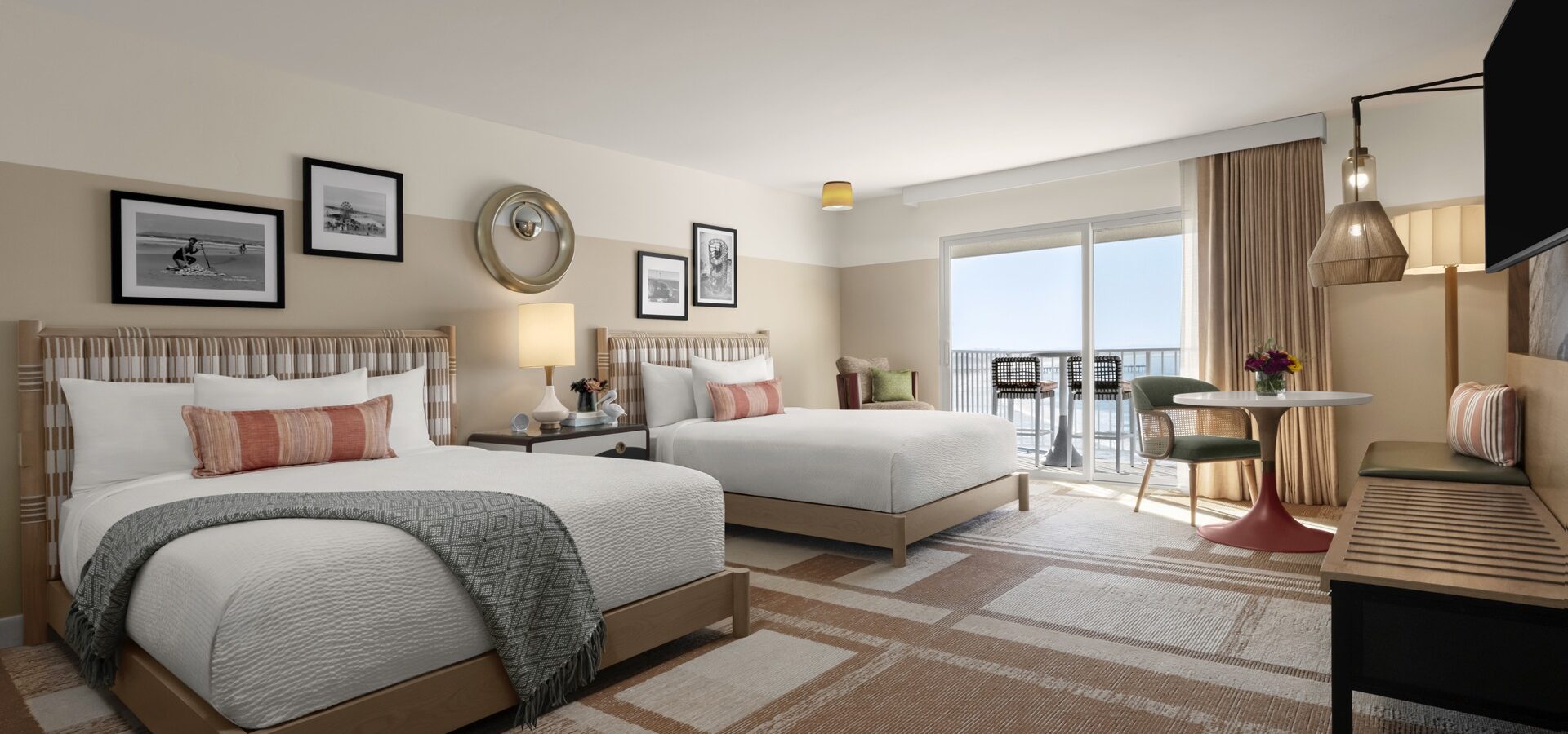 Modern hotel room with two double beds, decorative pillows, framed artwork on the walls, a round table with two chairs, a bench, and a large window with curtains leading to a balcony with an ocean view.