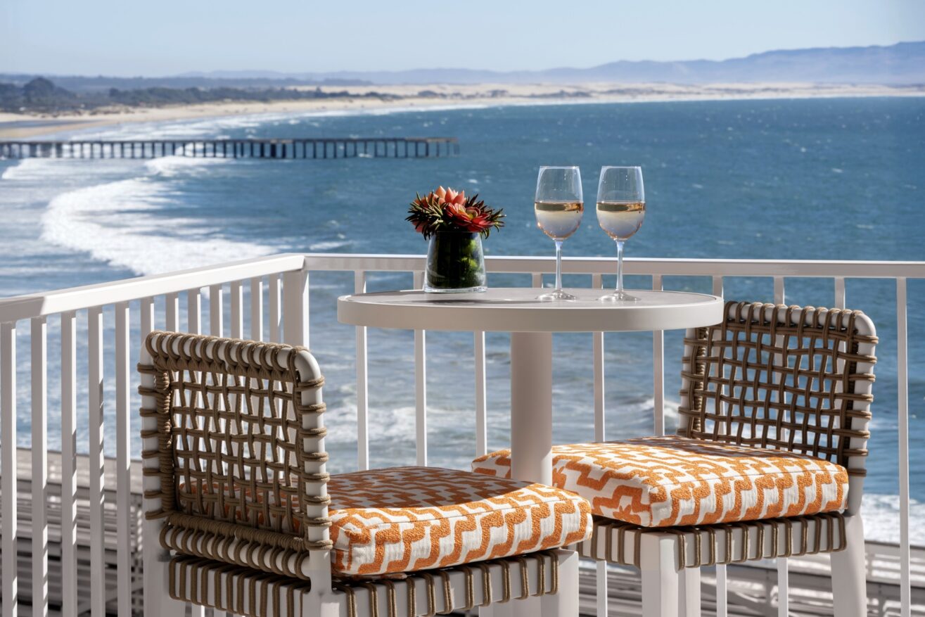 A small round table with two patterned chairs sits on a balcony overlooking the ocean. Two glasses of white wine and a small flower arrangement are on the table, with a scenic coastline and pier in the background.