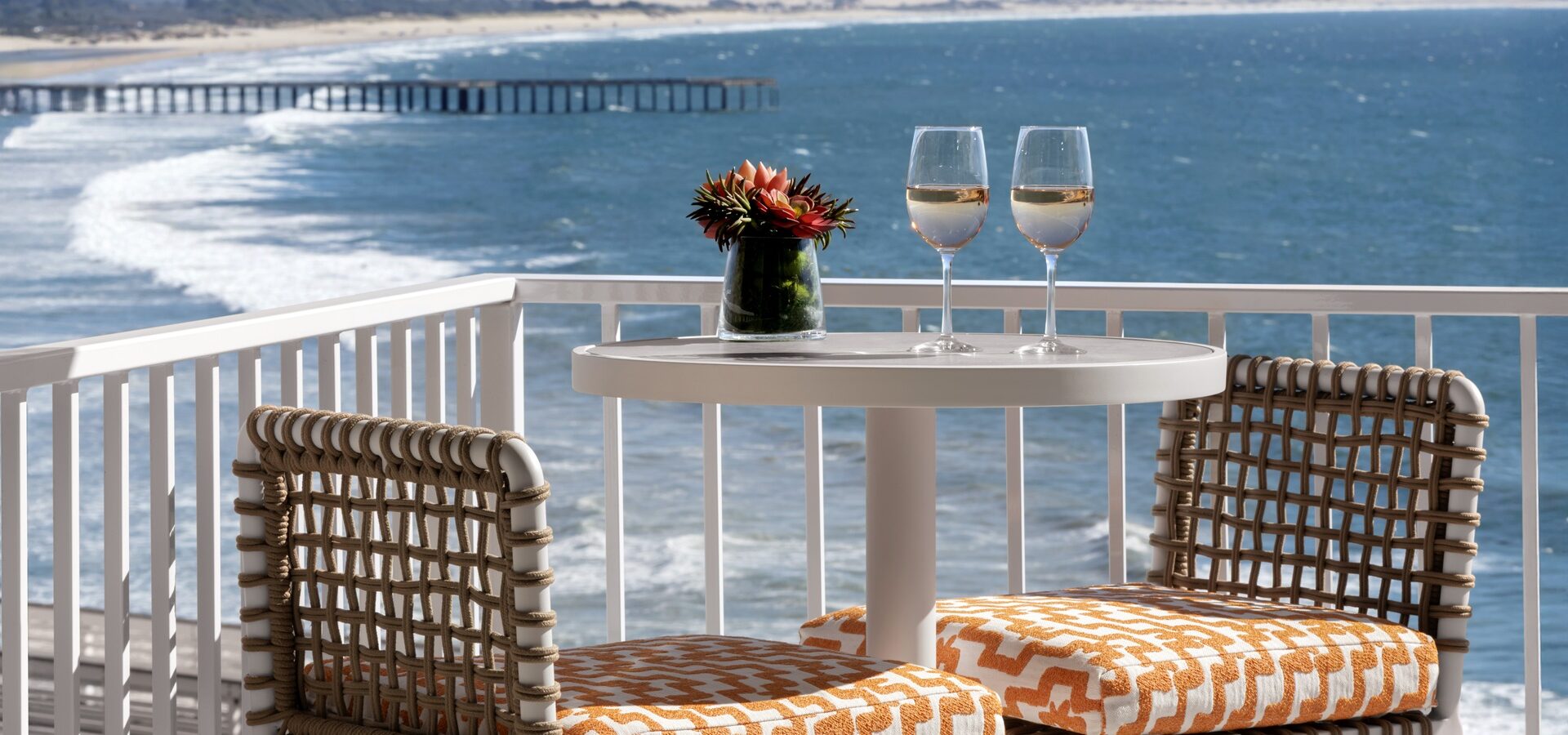 A small round table with two patterned chairs sits on a balcony overlooking the ocean. Two glasses of white wine and a small flower arrangement are on the table, with a scenic coastline and pier in the background.
