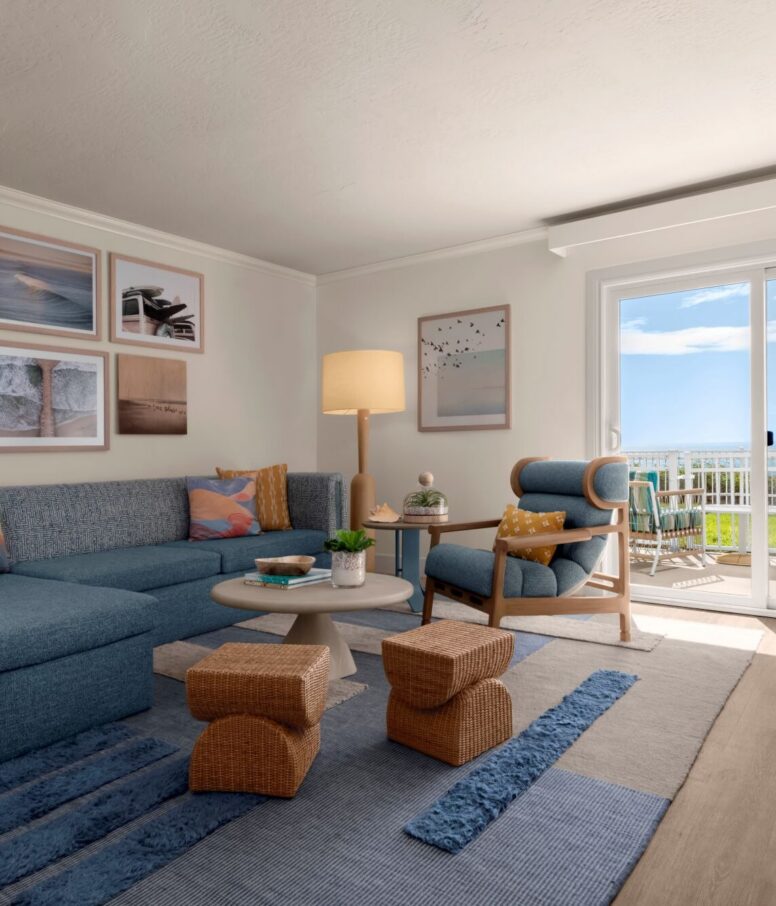 A bright living room with blue and beige decor, a sectional sofa, armchair, woven stools, coffee table, framed wall art, lamp, rug, and glass doors leading to a balcony with an ocean view.