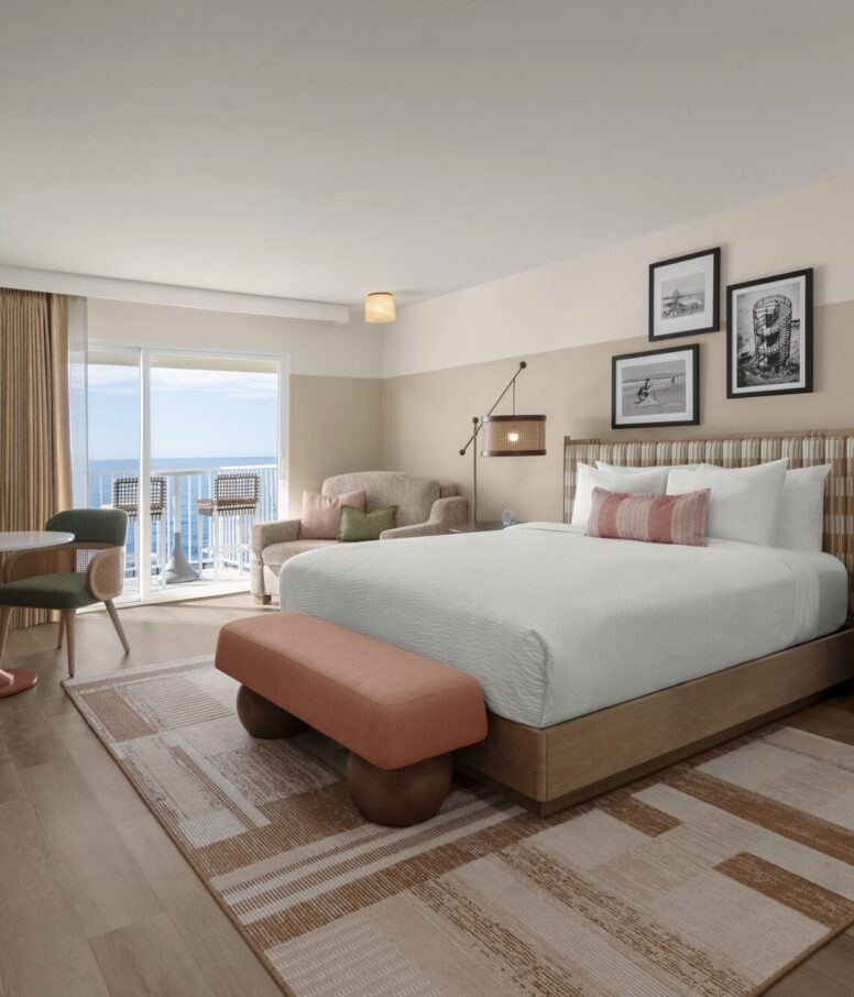 A modern hotel room with a large bed, peach and beige decor, a cozy armchair, wall art above the bed, a small dining area, and a balcony overlooking the ocean. Natural light fills the room.