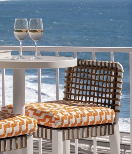 Two glasses of white wine sit on a round white table beside woven chairs with orange and white patterned cushions, overlooking a blue ocean with gentle waves.