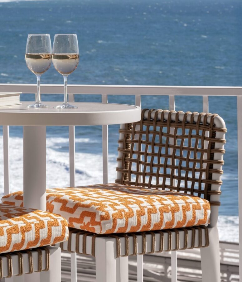 Two glasses of white wine sit on a round white table beside woven chairs with orange and white patterned cushions, overlooking a blue ocean with gentle waves.