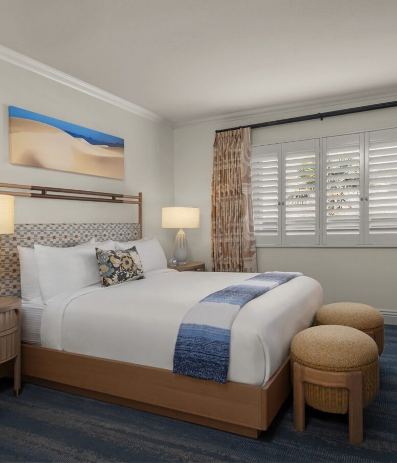 A neatly made hotel bed with white linens, blue accents, and floral pillows is flanked by two round ottomans. There are nightstands with lamps, a window with shutters, and a desert-themed painting on the wall.
