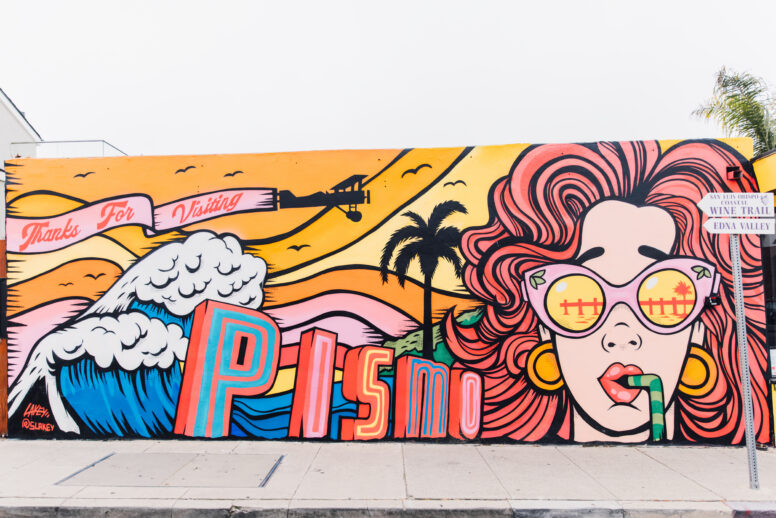 Colorful mural with waves, sunset, and a large face with red hair and sunglasses reflecting a pier. Text reads “Thanks for Visiting Pismo.” A palm tree and street sign are on the right side of the image.