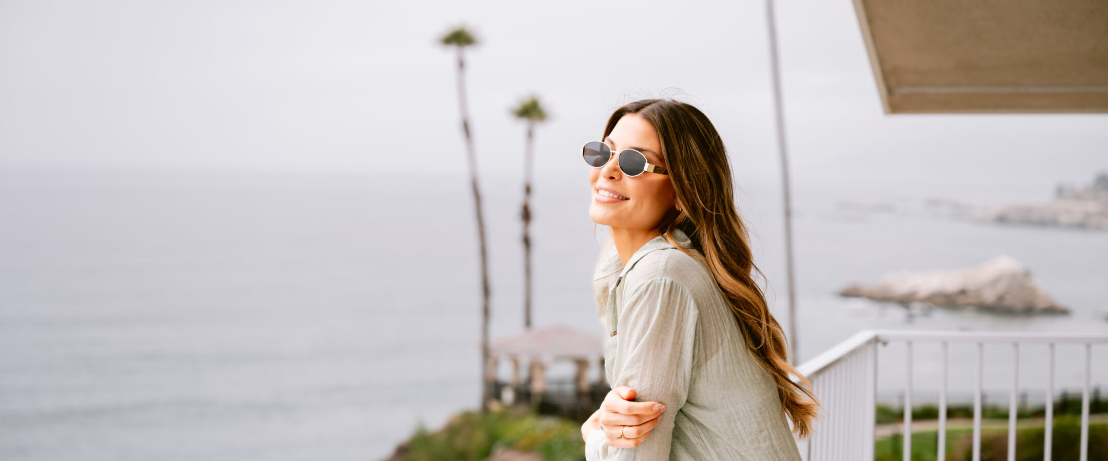 A woman in sunglasses and light clothing smiles while leaning on a balcony railing overlooking the ocean, with palm trees and a cloudy sky in the background.