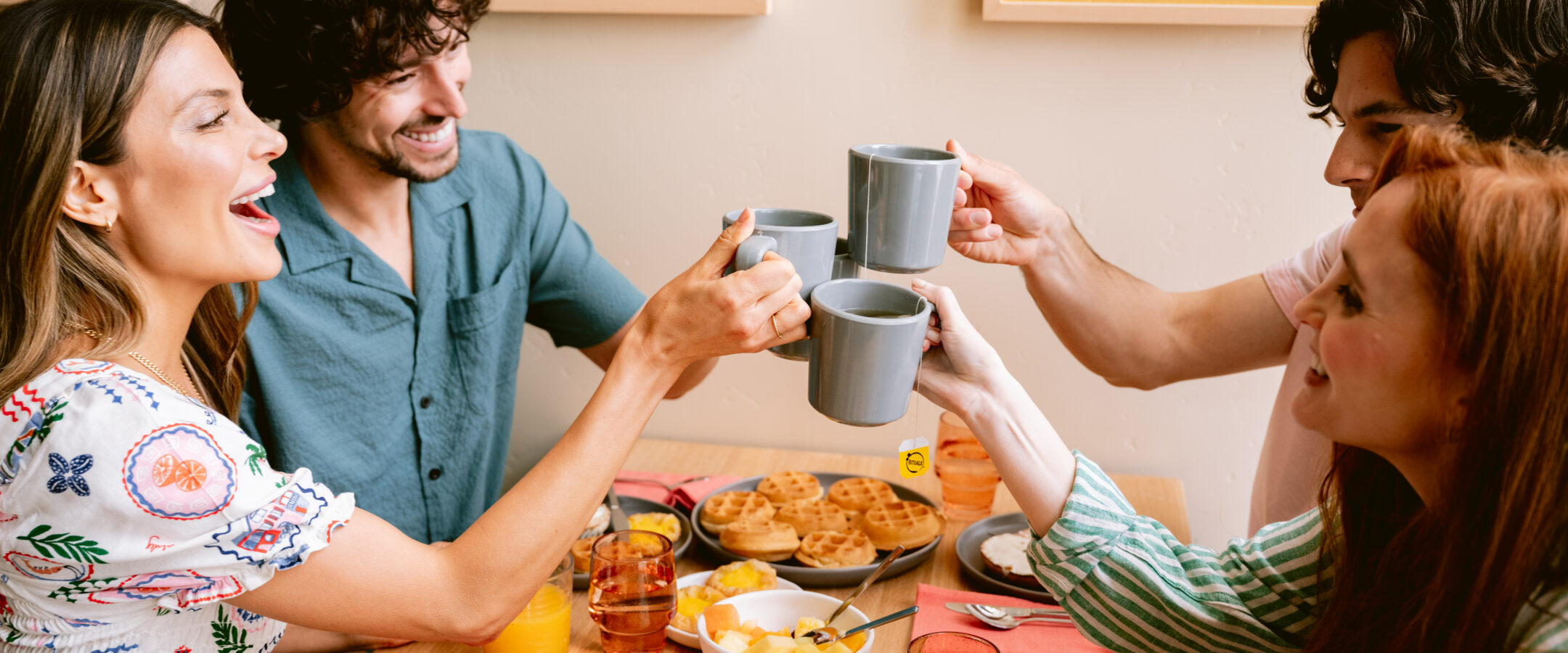 Four friends sit at a table, smiling and raising gray mugs in a toast over a breakfast spread of waffles, fruit, and orange juice. The mood is cheerful and casual.