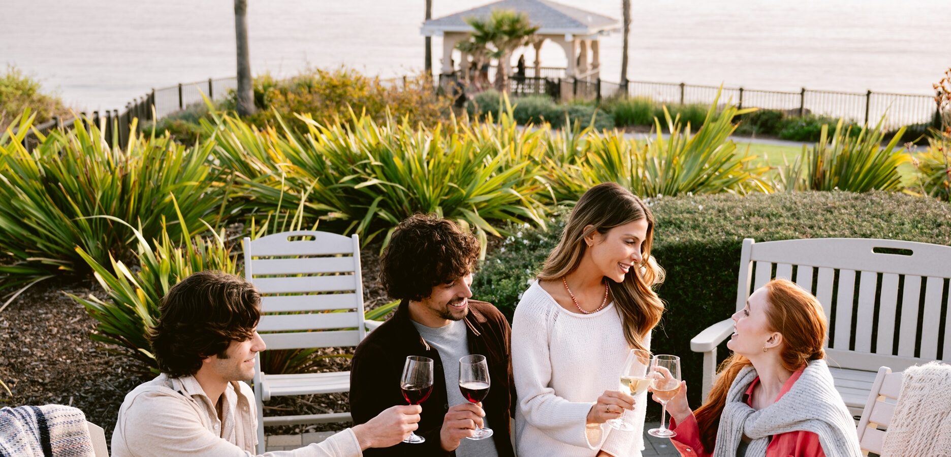 Four friends sit outdoors near greenery and toast with wine glasses, smiling and enjoying each others company. The ocean and a white gazebo are visible in the background.