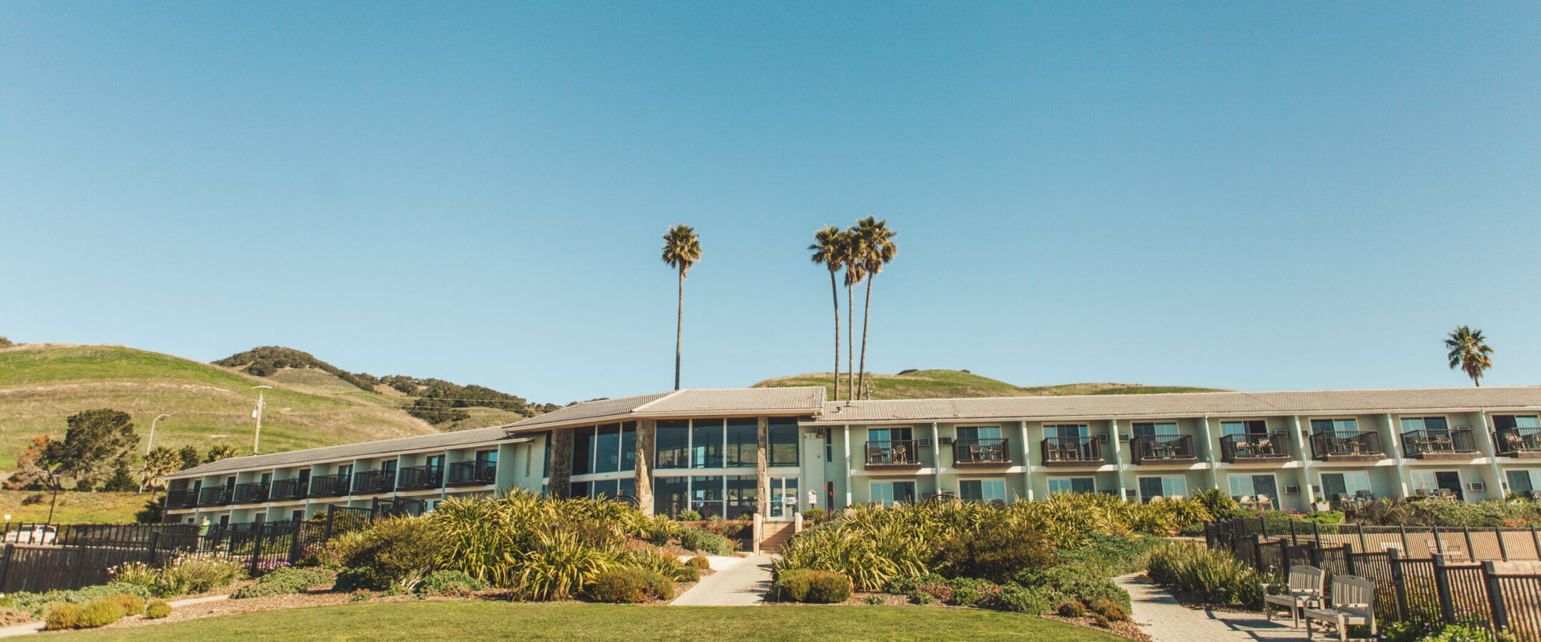 A modern, two-story hotel sits at the base of green hills under a clear blue sky, with palm trees rising behind it and landscaped gardens in front.